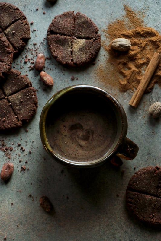 A mug of warm traditional Mexican hot chocolate and discs of chocolate de mesa with true cinnamon, whole nutmeg, and raw cacao beans.