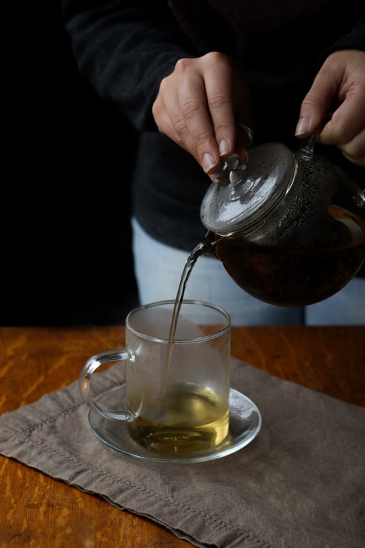 Midday herbal tea blend featuring lemon balm, holy basil, stevia, and schisandra berries. A glass tea pot dispenses a light golden tea into a clear glass mug for midday. 