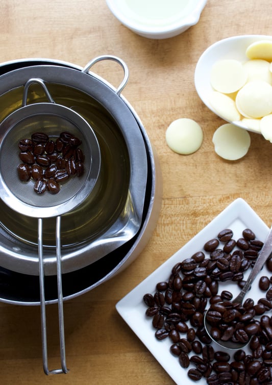 Strainer with coffee beans over a double boiler insert with dishes of cocoa butter wafers and coffee beans displayed around