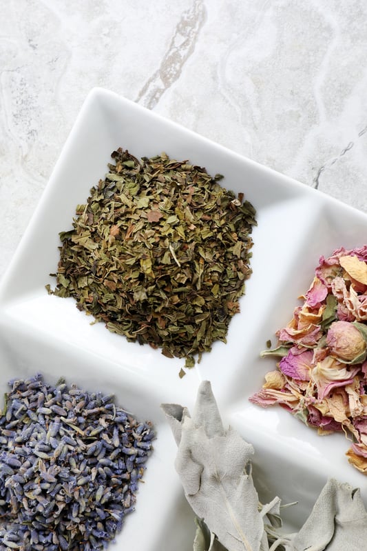 Dried herbs including lavender flowers and rose buds in divided white porcelain container on marble counter