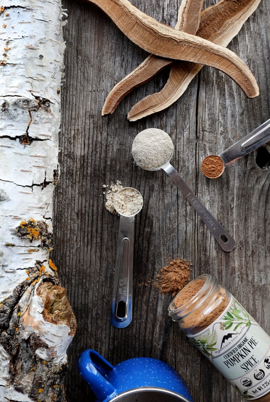 Spoons of colorful powder, reishi mushroom slices and a bottle of pumpkin spice spilling into rustic board. 