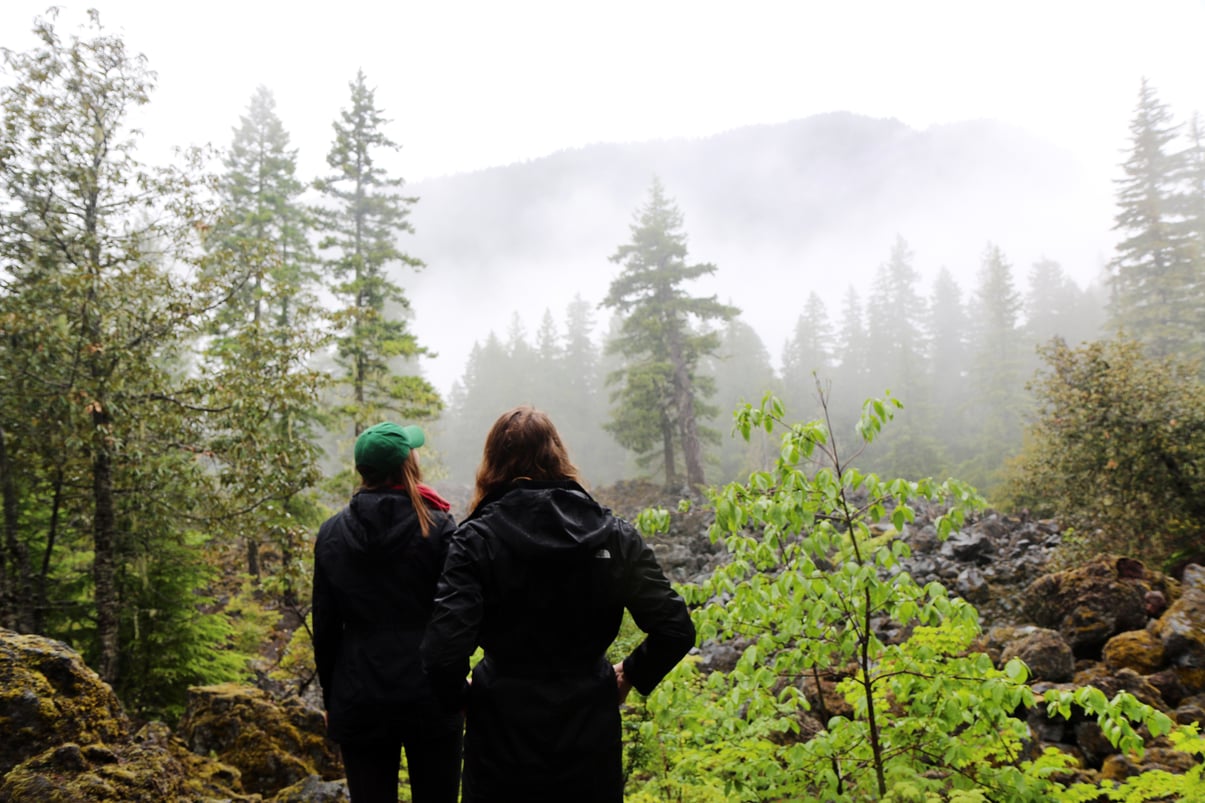 Two women pausing on a hike to look out upon a misty mountain view surrounded by evergreens.