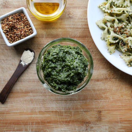 Nettle pesto in glass bowl near pasta and spoon with garlic powder, red pepper flakes, and olive oil