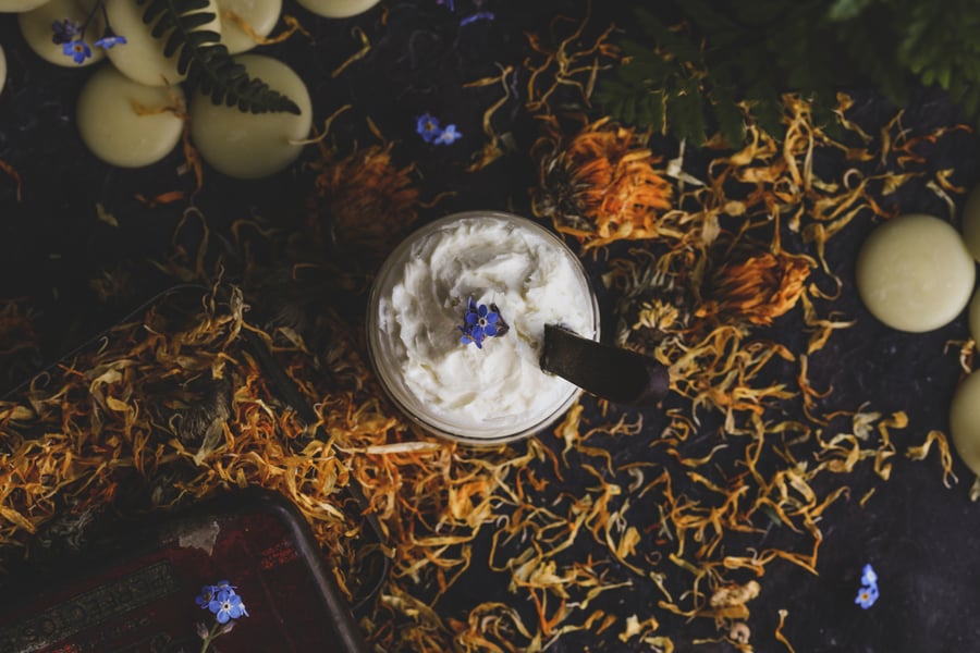 A jar of homemade nipple cream for breastfeeding sitting amidst calendula flowers and cocoa butter.