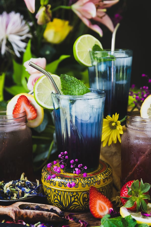 A colorful photo of four glasses of Nootropic Mocktails with focus on a blue glass of the Brain Rain Mocktail. The glasses are surrounded by bilberries, butterfly pea flowers, strawberries, and limes with out of focus florals in the background. The Brain Rain Mocktail is garnished with a metal straw, fresh mint, and a slice of lime.