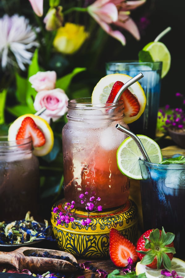A colorful photo of four glasses of Nootropic Mocktails with focus on a pink glass of the Lemon Love Berry Mocktail. The glasses are surrounded by bilberries, butterfly pea flowers, strawberries, and lemons with out of focus florals in the background.