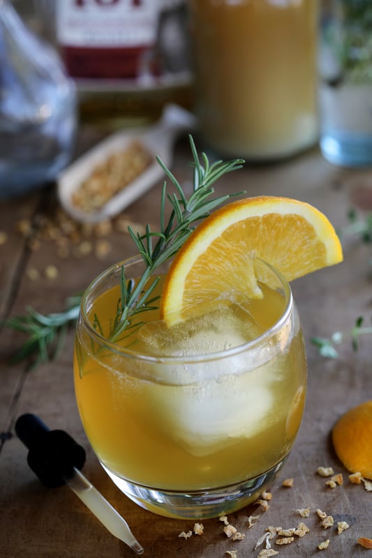 Small round glass with chilled orange color cocktail, fresh orange on rim and fresh rosemary sprig.