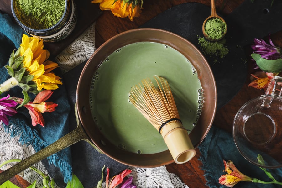 A bowl of matcha and a matcha whisk sit on a table surrounded by flowers and a jar of matcha powder.