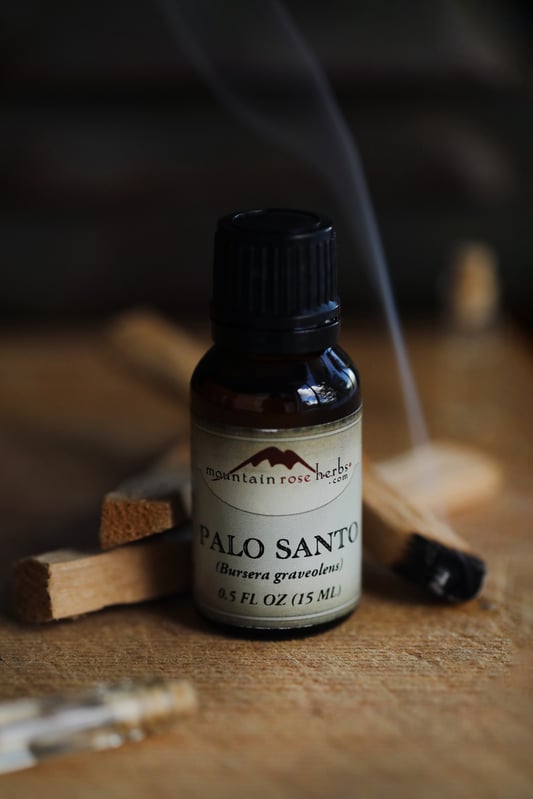 A half-ounce bottle of palo santo essential oil flanked by burning pieces of palo santo. Small ribbon of smoke is coming up from the burning end of a palo santo stick.