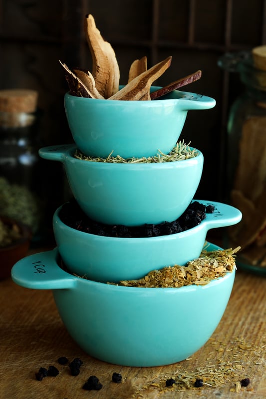 Bright sky blue measuring cups are full of herbal ingredients and stacked high. Reishi mushrooms, rosemary, and bilberries fill the measuring cups which are used to add ingredients in parts to herbal recipes. 