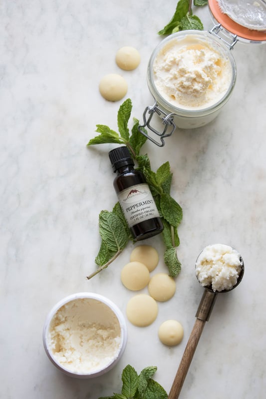 Bottle of essential oil displayed with fresh mint, cocoa butter wafers and whipped body butter