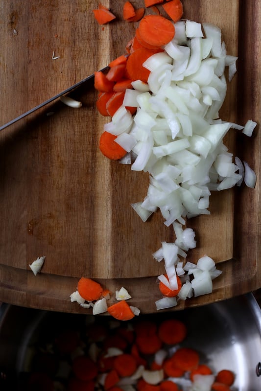 Wooden cutting board with knife actively dicing carrots, onions and garlic. 