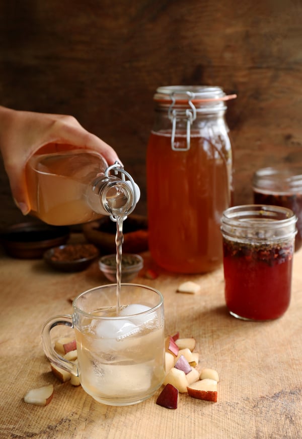 Pouring flavored kombucha into an iced mug. Jars of kombucha are fermenting in the background and fresh pears and herbs add flavor to the beverage. 
