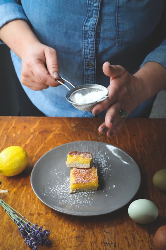Hands using strainer to dust lemon bars with powdered sugar surrounded with fresh ingredients