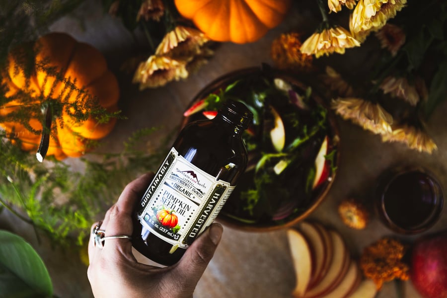 A bottle of Mountain Rose Herbs Pumpkin Seed Oil is being poured from a hand into a bowl of salad surrounded by fall items like small pumpkins and orange flowers.