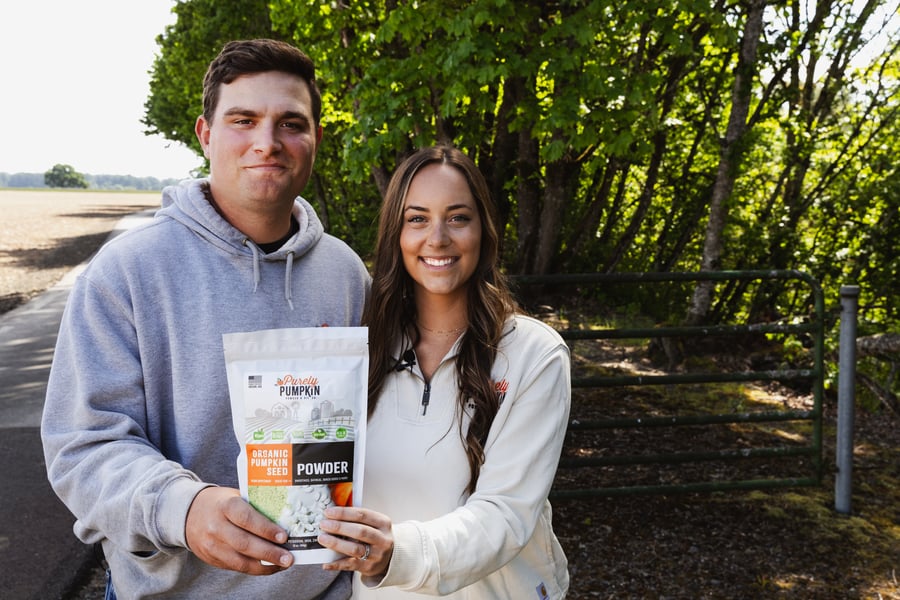 Two pumpkin farmers stand proudly in front of their fields showing one of their many products, a bag of their organic pumpkin seed powder.