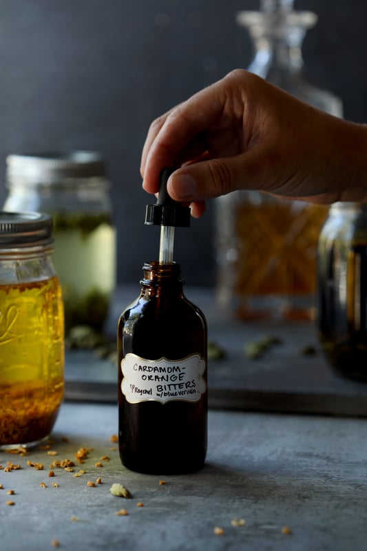 Homemade bitters featuring cardamom, bitter orange peel, and blue vervain in an amber dropper bottle with herbs tincturing in the background.