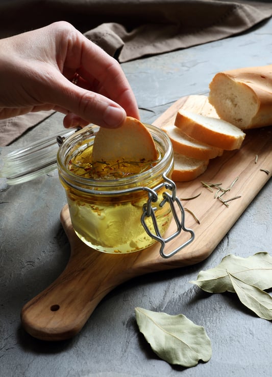 Hand dipping bread in jar with herb marinated goat cheese with cutting board on counter and loaf of bread