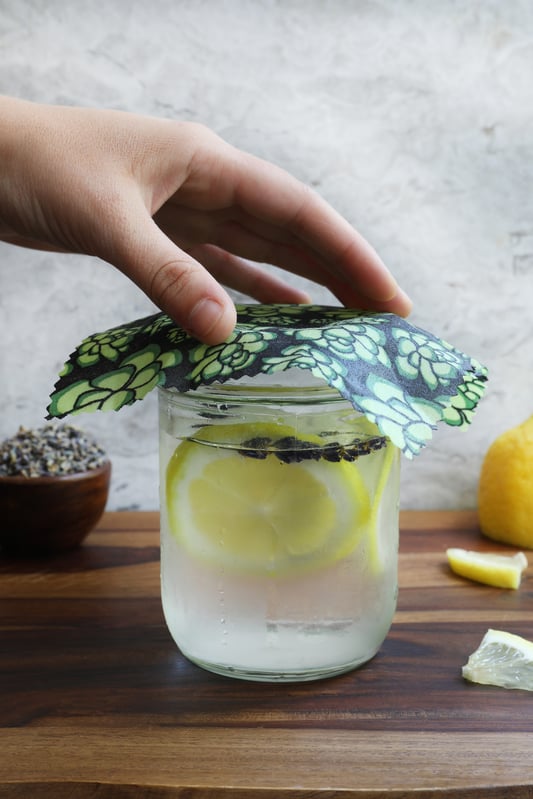Hand placing colorful beeswax wrap on top of glass of iced water with sliced lemon and botanicals. 
