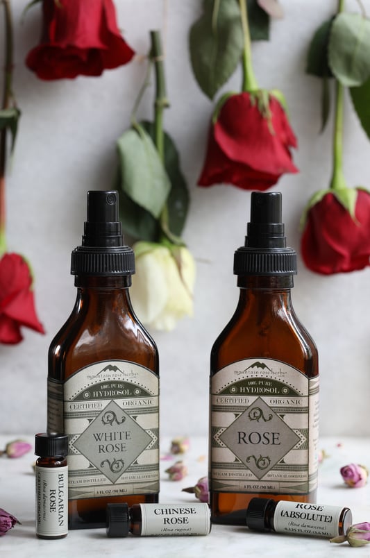 White rose hydrosol, rose hydrosol, and various rose essential oils laying out on counter with rose petals