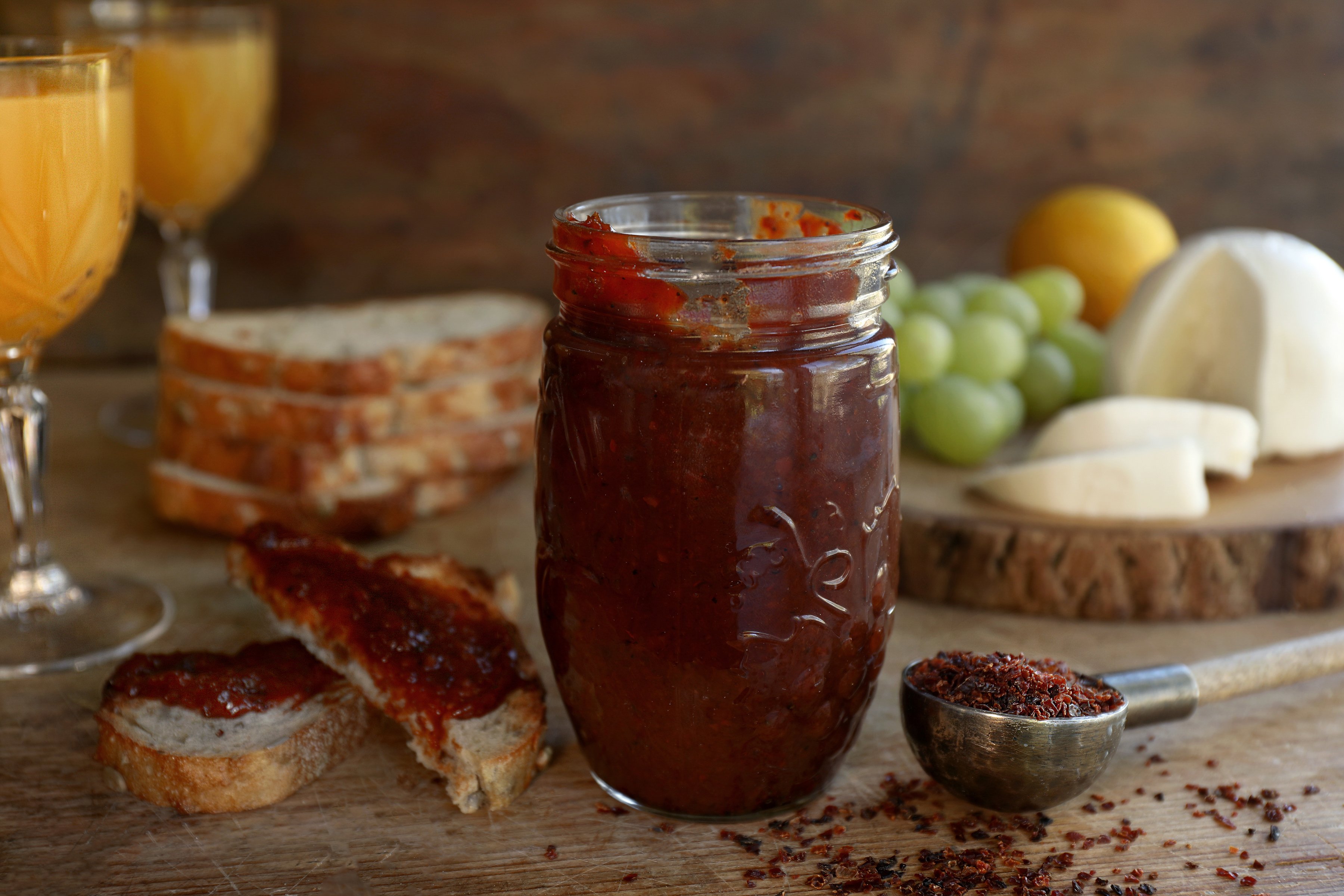 A jar of homemade rosehip jam using dried rosehips and other ingredients. Toasted whole grain bread and a cheese and fruit board with orange juice in the background.