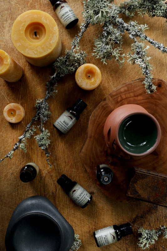 Aromatherapy products, including beeswax candles, terra cotta diffusers, and essential oils, are assembled on a wooden table with lichen-covered branches.
