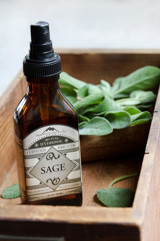 Amber mister bottle of organic sage hydrosol in shallow wooden crate with fresh sage leaves