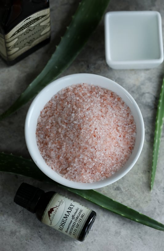 A bowl of granulated pink Himalayan salt placed in the middle of fresh aloe vera leaves, a bottle of rosemary essential oil, and peppermint hydrosol.