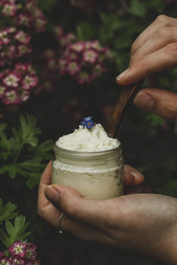 Homemade whipped skin cream in clear jar with purple flowers on top. 