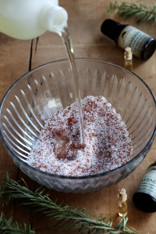Pouring oil into bowl of colorful salts with vials of essential oils displayed around bowl. 