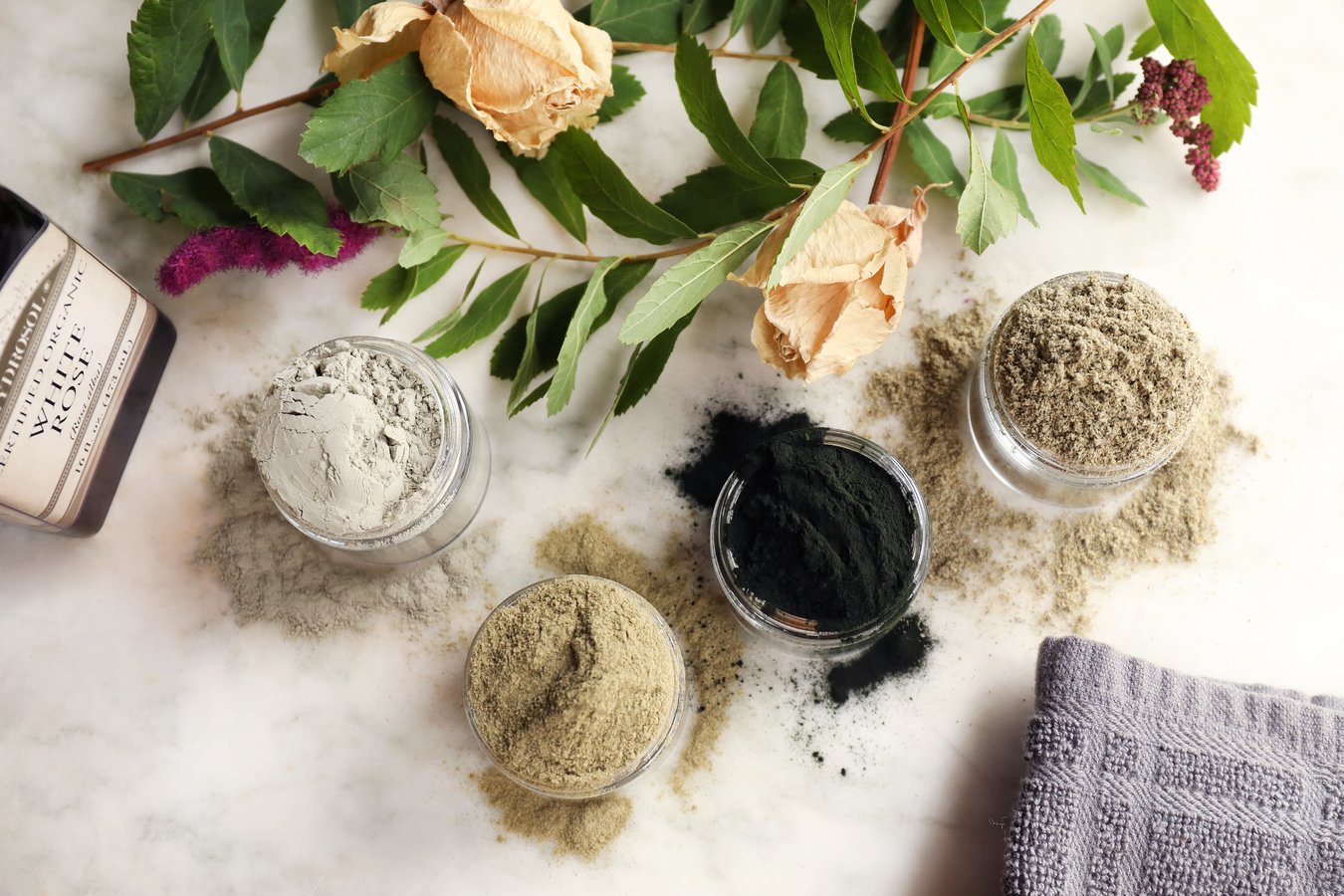 A variety of clay, spirulina, and seaweed powders in jars  for dry face mask recipe, set among roses and bay branches, with Mountain Rose Herbs bottle of rose hydrosol to add liquid to mask base.