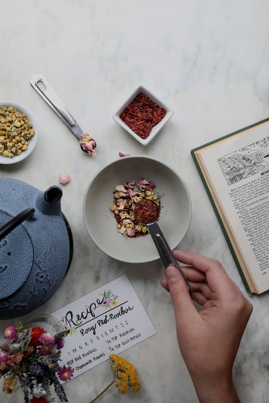 Hand holding spoon of rooibos tea blend over bowl in order to mix herbal ingredients into a tea