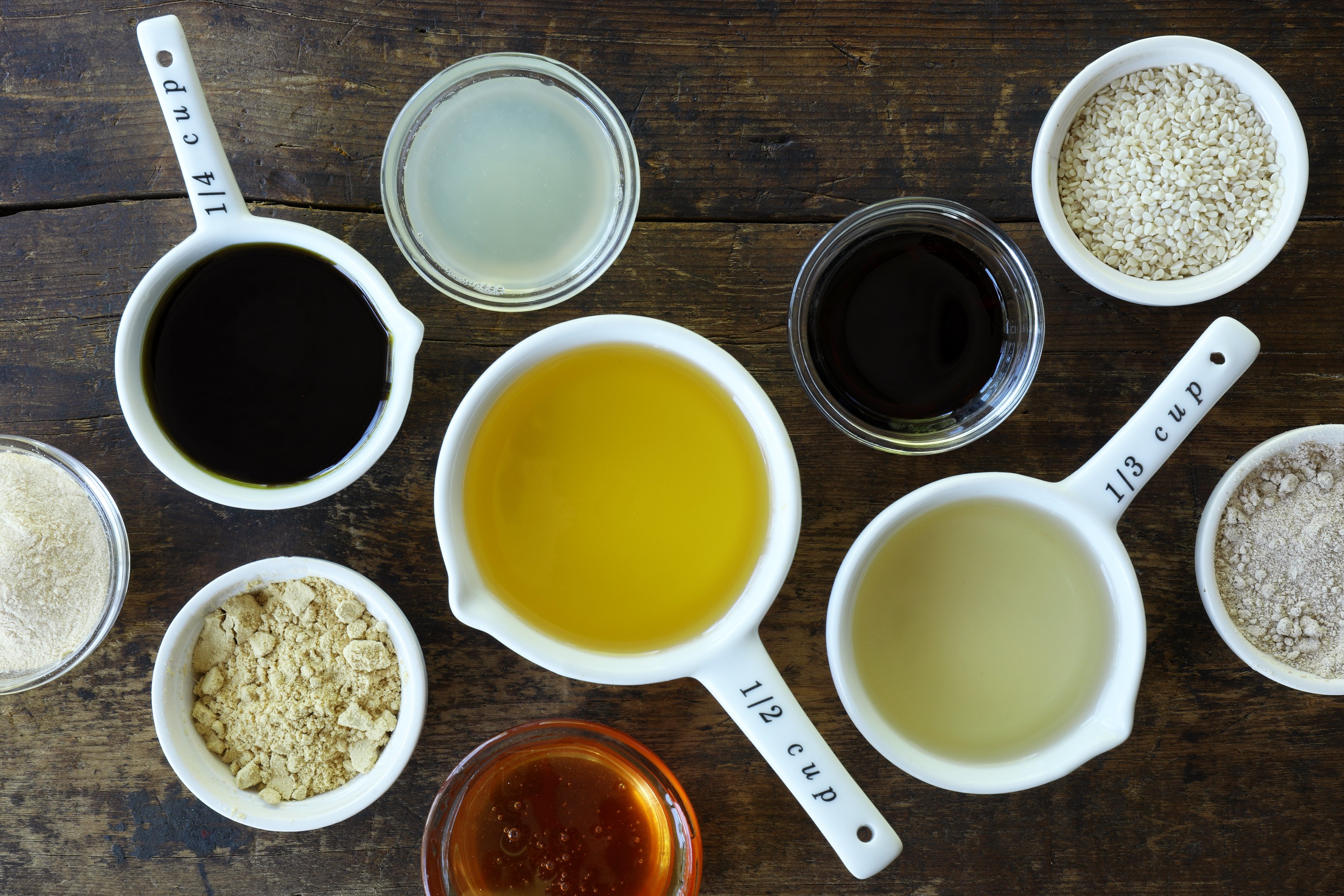 Measuring cups and bowls of oils and spices for Asian salad dressing arrayed on wooden table.