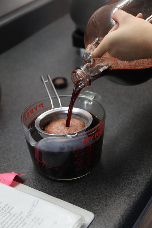 With a recipe in sight, a solution of elderberry syrup is poured from a large glass jug through a strainer into a measuring bowl. 
