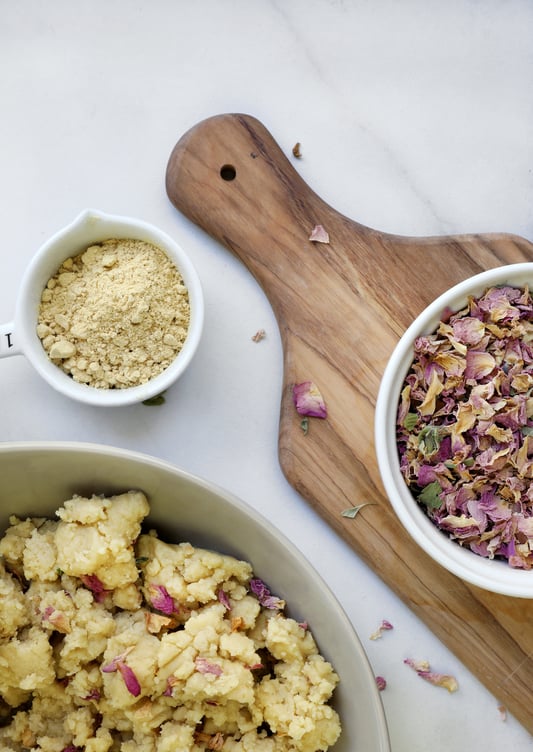 Rose petals in bowl sitting on wooden cutting board near a ceramic cup full of flour and a large mixing bowl of dough 