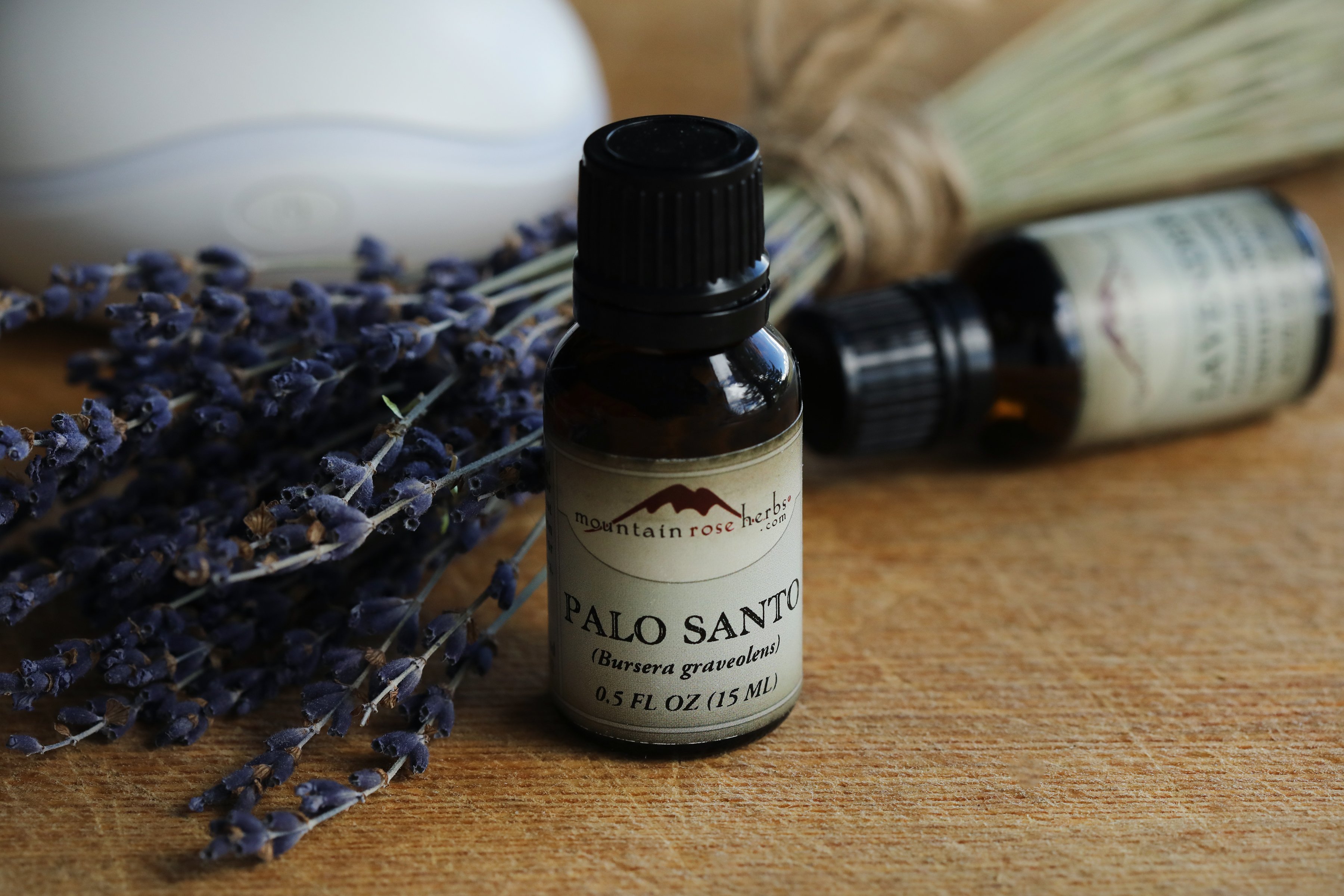 Palo santo and lavender essential oils arranged with a bouquet of lavender flowers. An aromatic diffuser is in the background.  