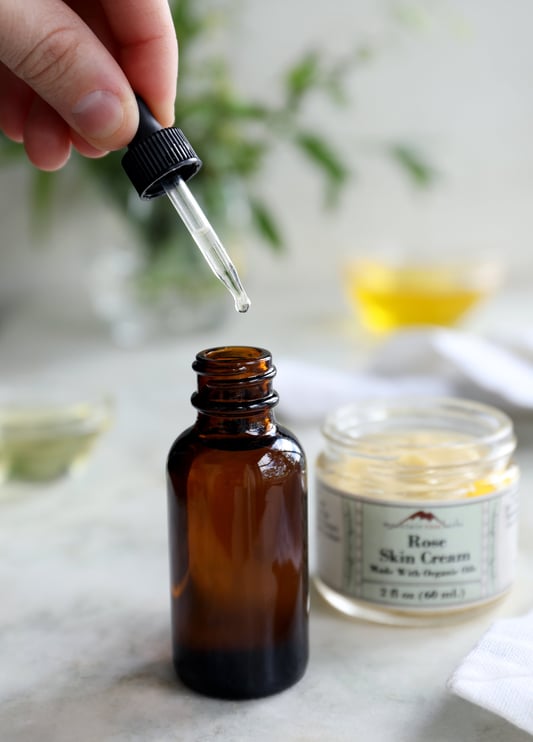 An amber glass bottle with a glass dropper pulling out an oil formula with a jar of rose skin cream in the background. 