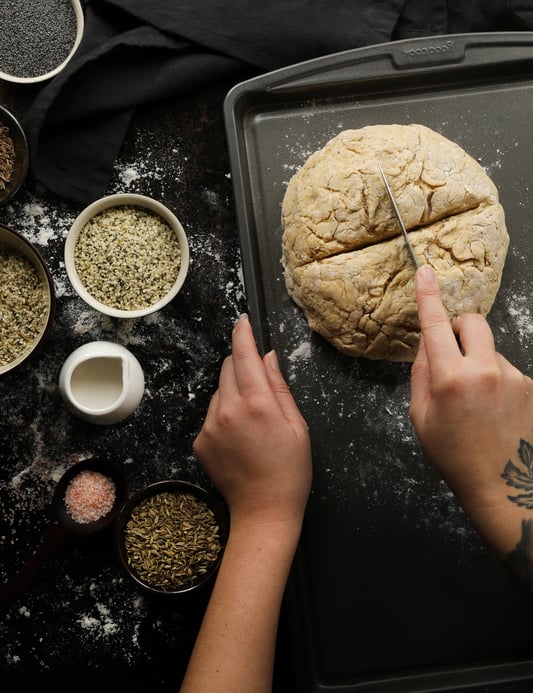 Scoring a loaf of Irish soda break that is ready to be put in the oven to bake. Made with nutritional seeds like hulled hemp seeds, poppy seeds, caraway seeds, and fennel seeds, this crunchy soda bread is great for St. Patrick's Day.