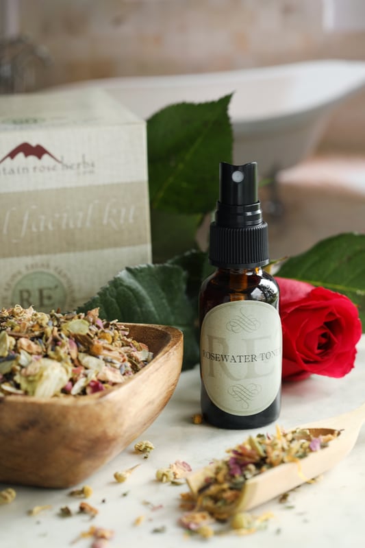 Relaxing herbs arranged in a wooden bowl for a spa treatment. A red rose sits behind a bottle of rose water, and a wooden scoop spills over with dried herbs.