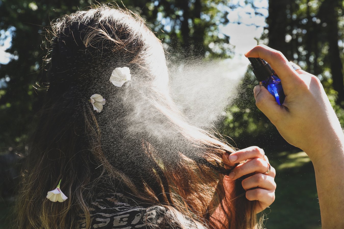 A woman mists her hair with DIY salt spray in the sunshine, there are flowers in her hair.