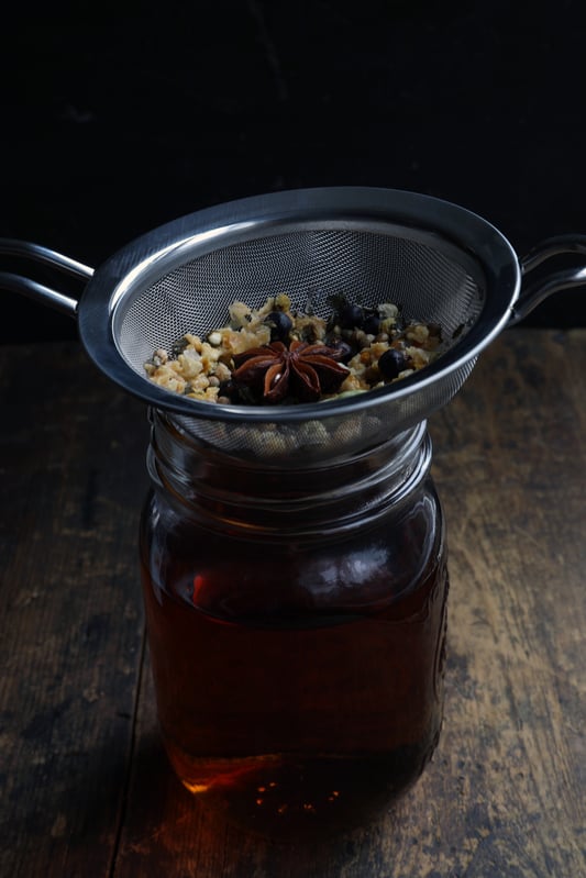 Strainer holding boiled herbs and spices over glass pantry jar with strained vermouth