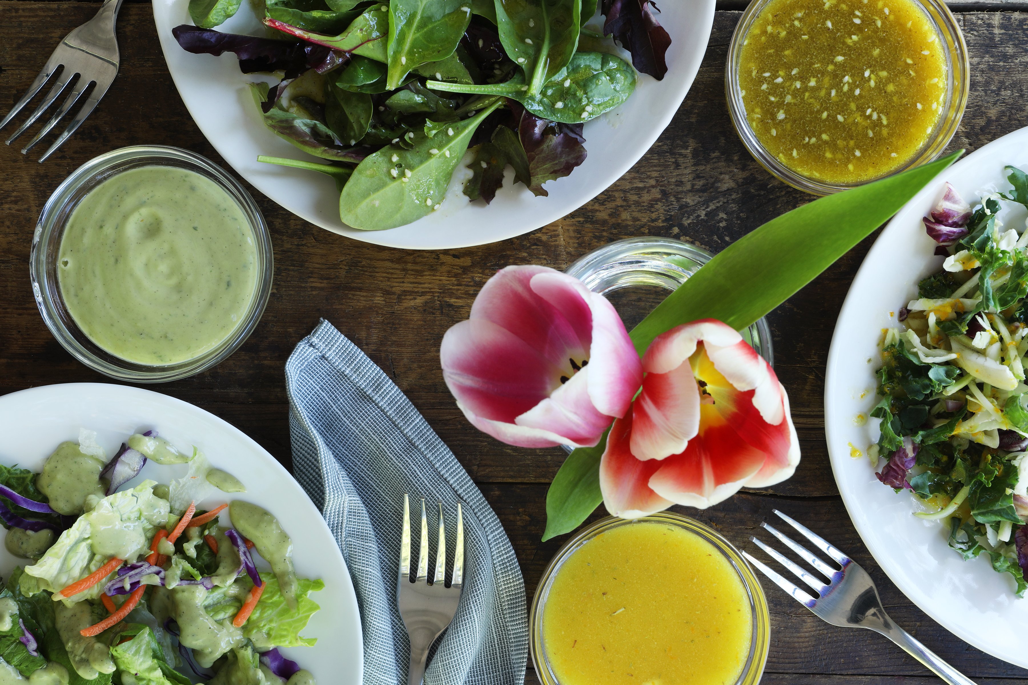 Three bowls of fresh salad greens arranged with fresh tulips and organic salad dressings.