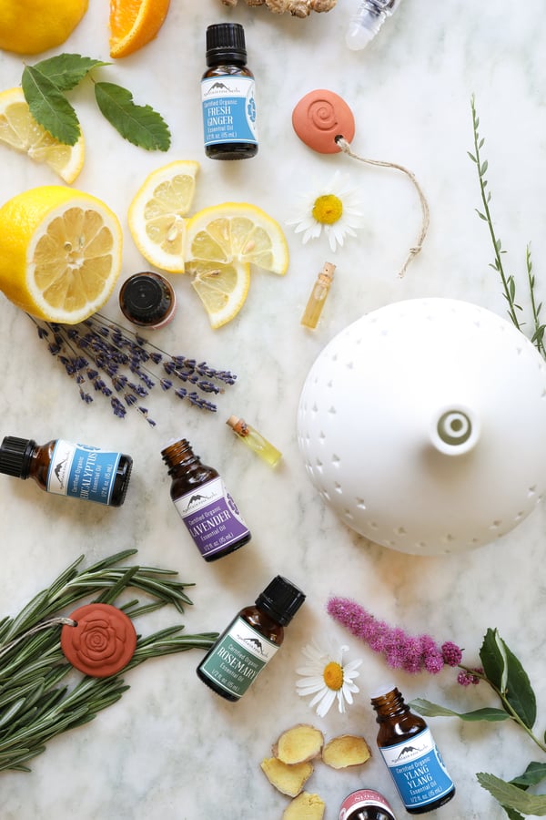 Two varieties of ceramic essential oil diffusers surrounded by essential oil bottles and fresh botanicals.