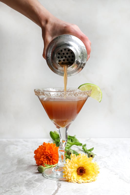 Hand pouring herbal cocktail into margarita glass with smoked salt on rim and flowers on counter