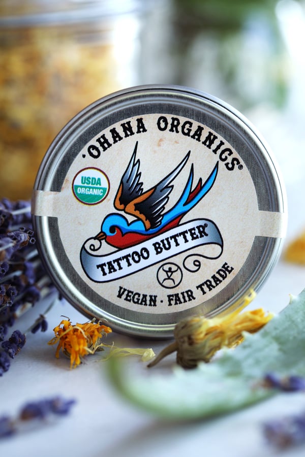 Product photo of Ohana Organics tattoo butter. All-natural and vegan tattoo after care salves arranged with freshly dried herbs such as lavender, calendula, and aloe vera.