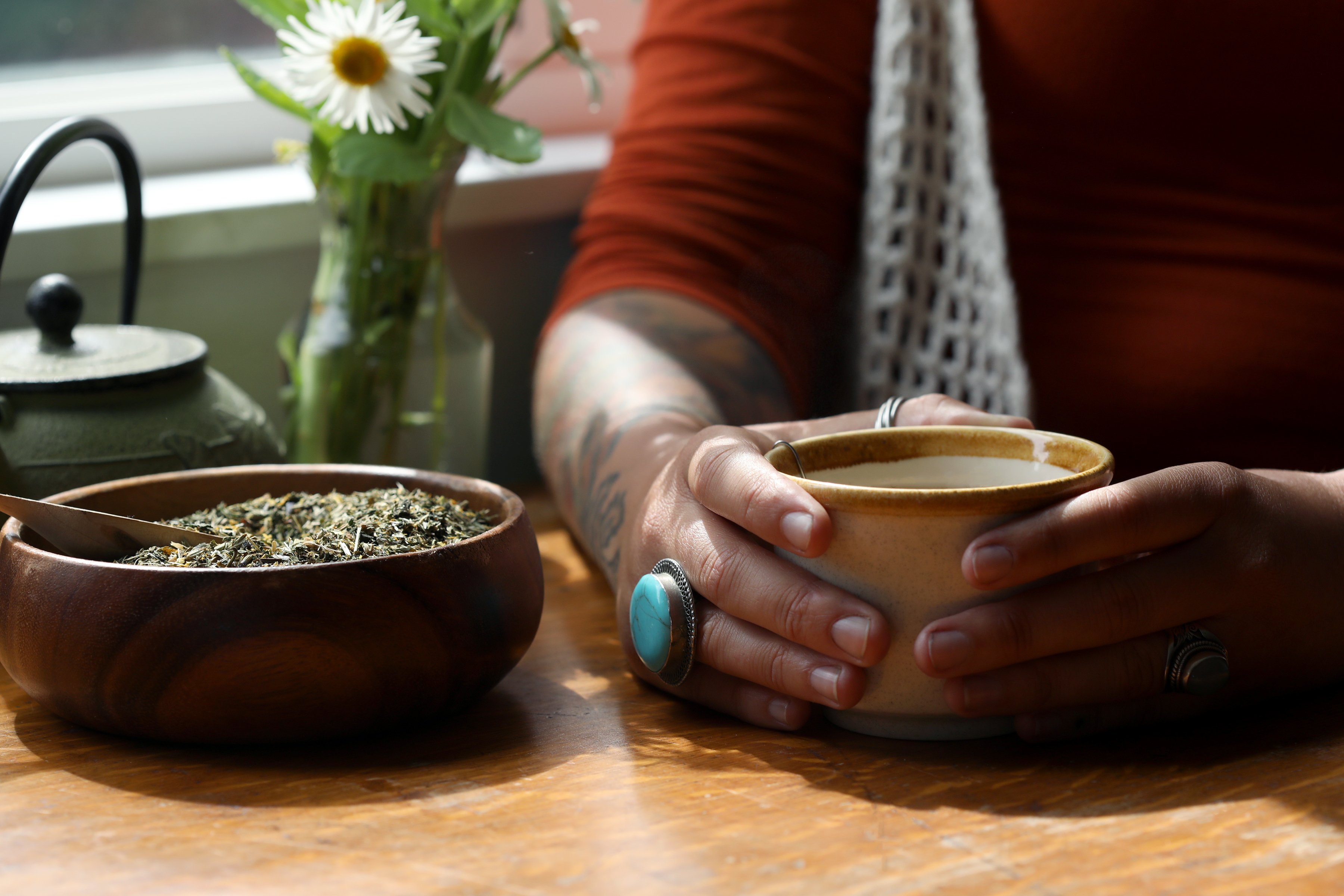 Tattoo care by drinking nettle and blossoms herbal tea. A young woman sitting with a bowl of herbs and a fresh cup of tea next to a window sill with cut flowers in bloom.