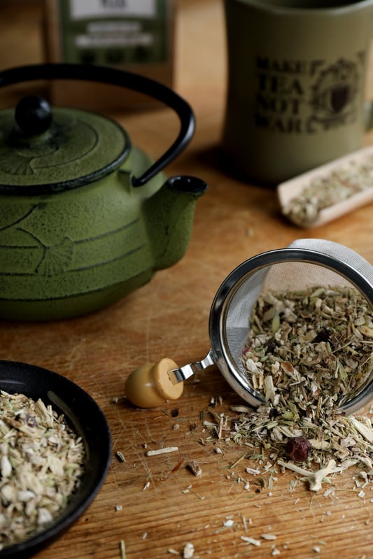 A collection of tea accessories perfect for Father's Day gifts. A small tea strainer with a wooden handle, wooden scoops, a green ceramic tea pot, and green mug. A blend of dried herbs spilling from the strainer. 