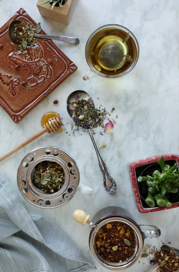 Various loose leaf teas are being steeped in mugs using different infusers. A mesh tea ball, and mesh strainers are full with loose herbal teas. Honey stick and vintage spoon are overflowing. 