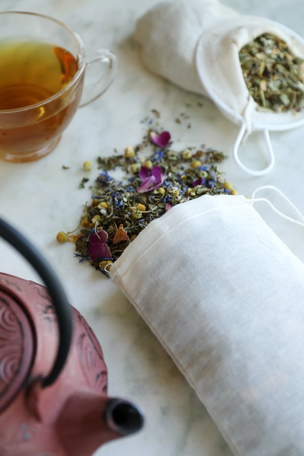 A cotton tea sock and cotton muslin bag are both filled with loose leaf herbal teas. A brewed cup of tea and a stainless steel tea pot are arranged nearby. Colorful herbal tea blend of rose petals, chamomile, lavender, corn flowers. 