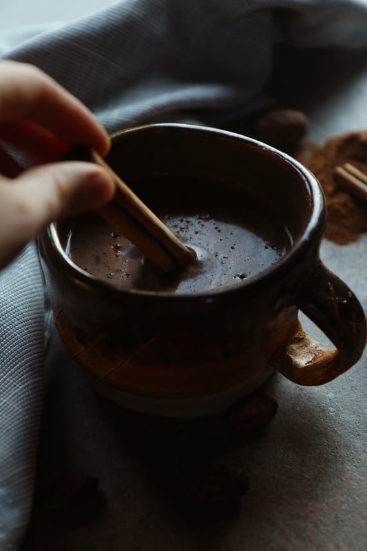 A mug of warm traditional Mexican hot chocolate being stirred with a stick of Ceylon cinnamon. Mexican hot chocolate is rich, dark, creamy, and warming during cold days. 